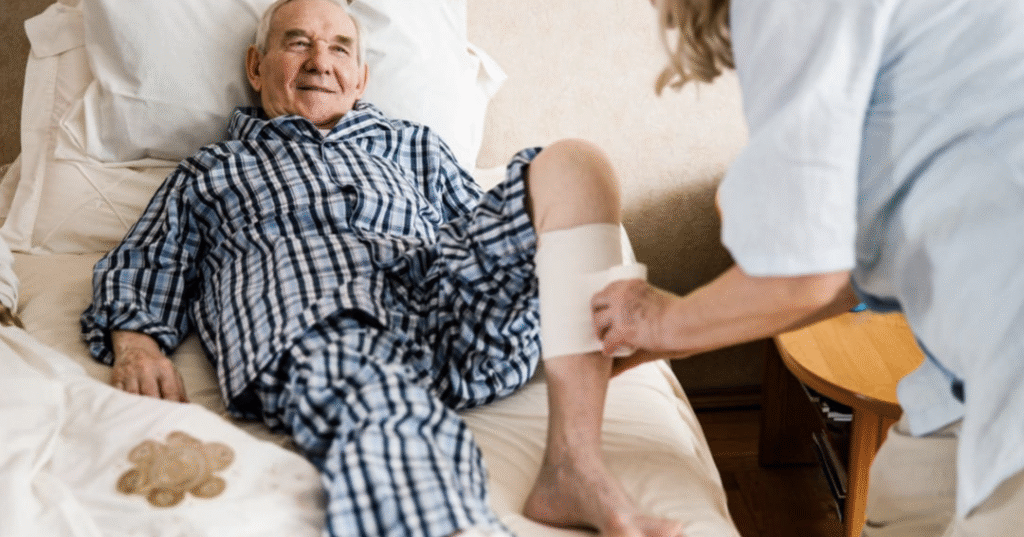 An older man in checkered pajamas lies in bed while a healthcare professional applies a bandage to his lower leg.